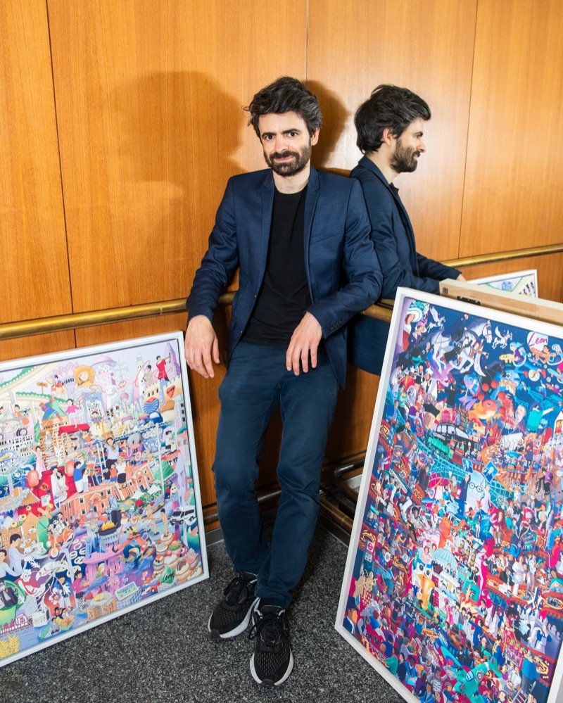 Leon Fenster leaning on a rail between two of his framed, colourful city artworks